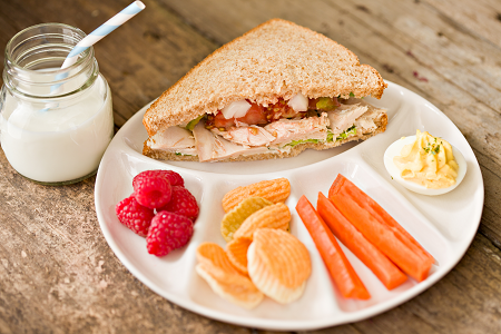 A white plate with separate sections sits on top of a wooden table. The plate has each section filled, one with a sandwich and the others filled with fruits and veggies. A small glass of milk with a straw sits next to the plate