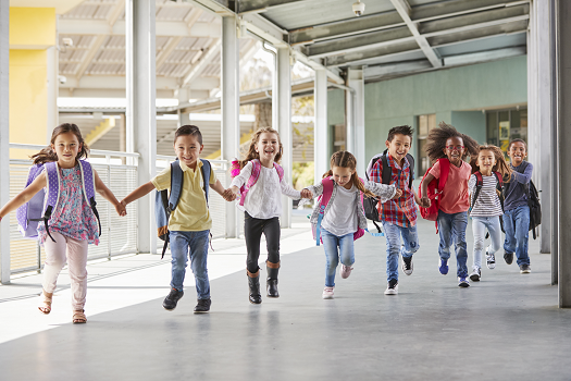 A group of cheerful children with backpacks run hand in hand down a covered walkway at a school. The kids, who are diverse in appearance and wearing colorful clothing, are smiling and appear to be excited, possibly about recess or the end of the school day.