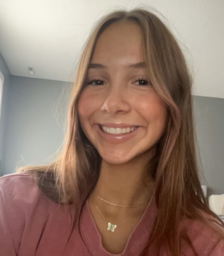 Addie, a young woman with long brown hair, smiles warmly at the camera. She's wearing a pink shirt and a necklace with a butterfly pendant. The softly lit room with grey walls in the background adds to the gentle ambiance of the scene.