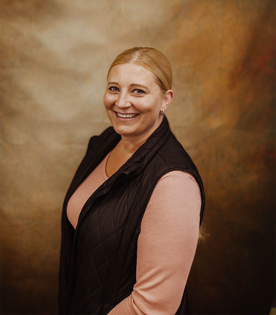 Amanda with blonde hair smiles at the camera against a warm, brown textured background. They are wearing a pink top and a black quilted vest.