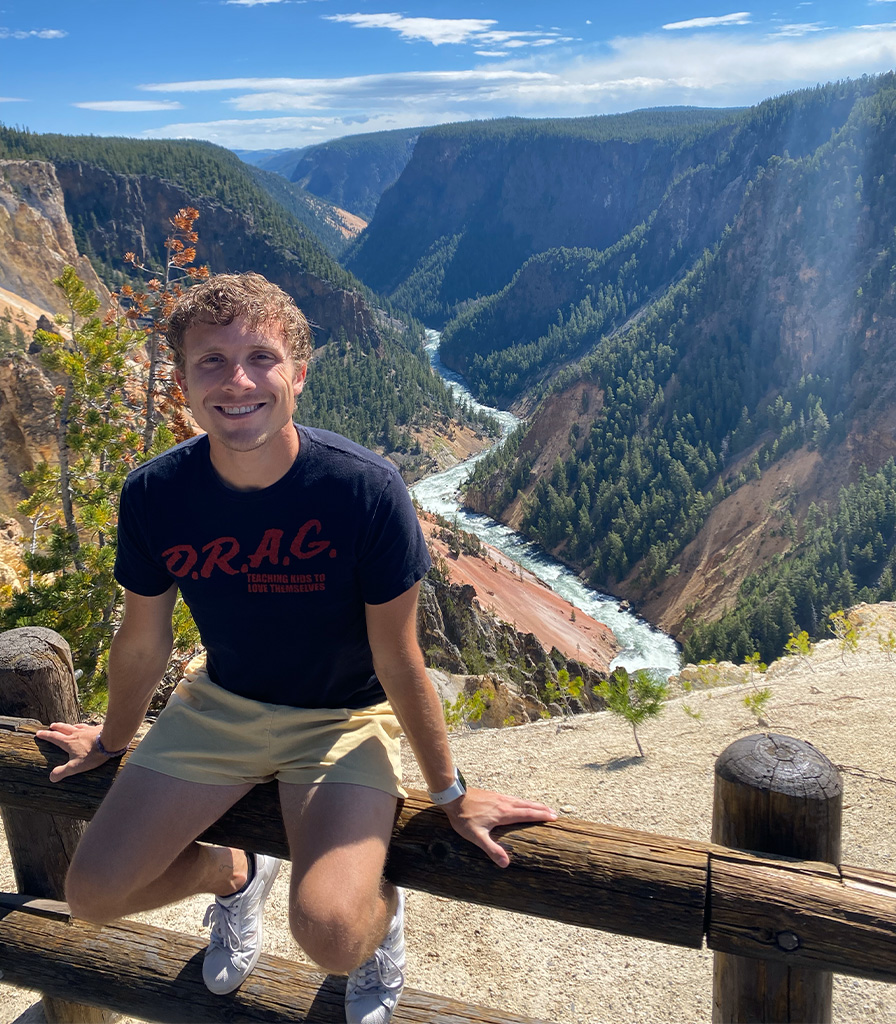 Austin smiles while sitting on a wooden fence overlooking a scenic canyon with a river flowing through it. The background features steep cliffs, lush greenery, and a clear blue sky.