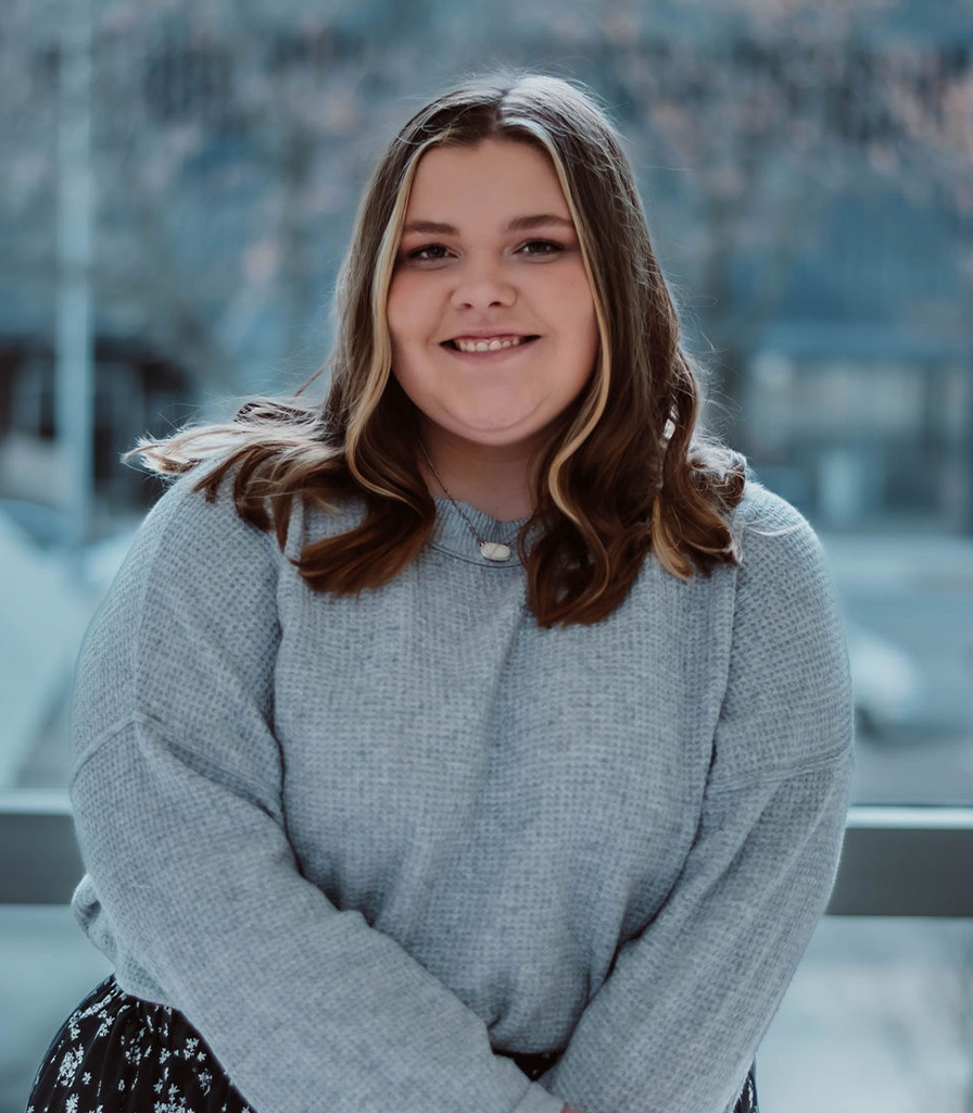 Callie, with long brown hair and a cozy gray sweater, smiles warmly while sitting indoors. Behind her, a large window offers an out-of-focus view of the bustling cityscape.