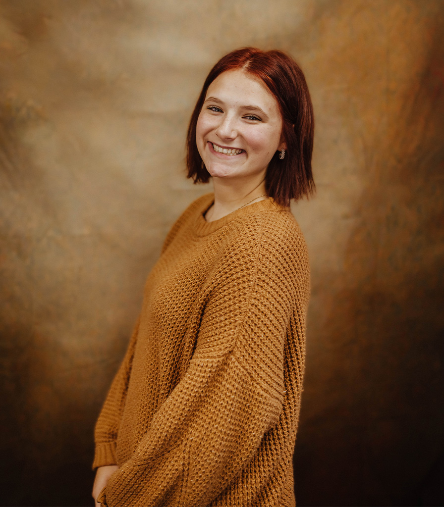 Cassie, with shoulder-length reddish-brown hair, is smiling warmly in a textured brown sweater. The background offers a golden-brown hue, creating a soft and cheerful ambiance to the portrait.