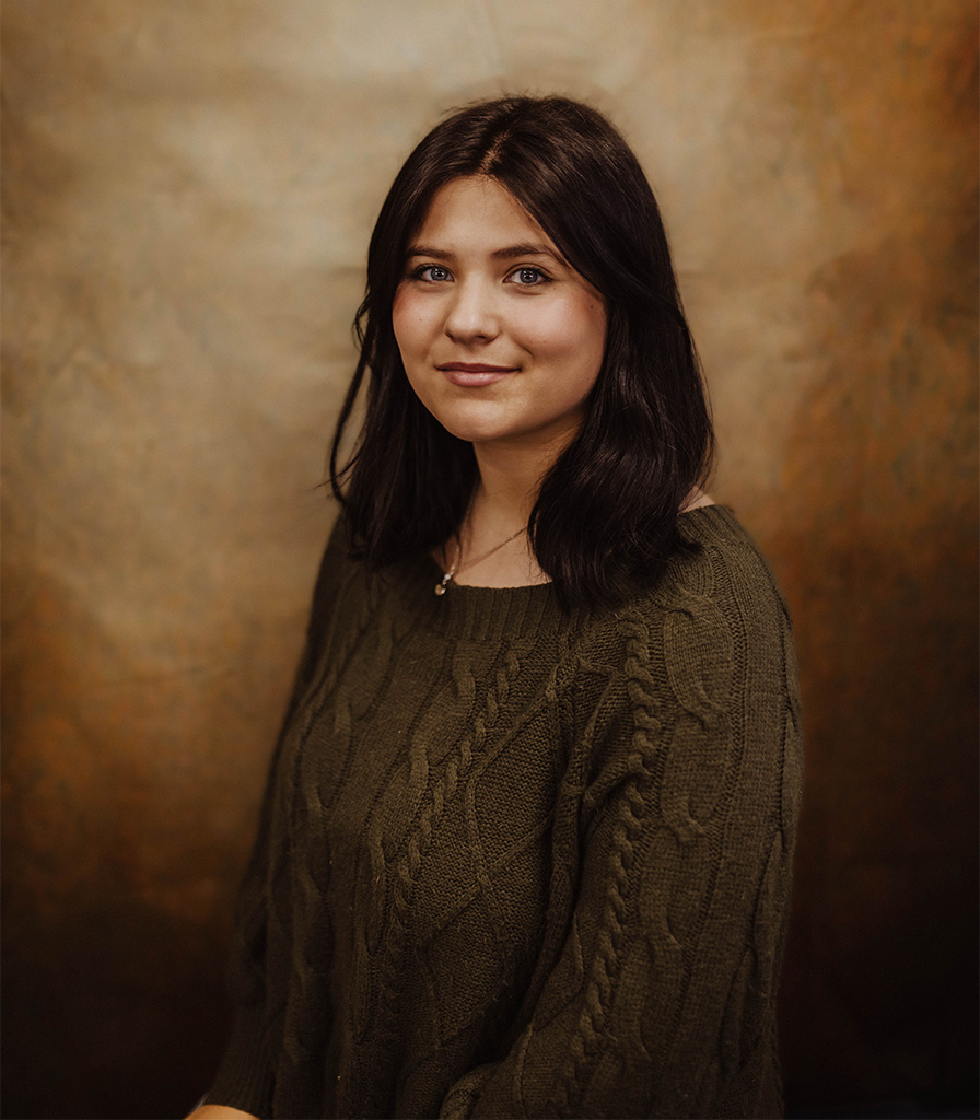 Codi, with shoulder-length dark hair, sits comfortably in a green cable-knit sweater against a warm, textured brown background. They look forward, smiling softly.