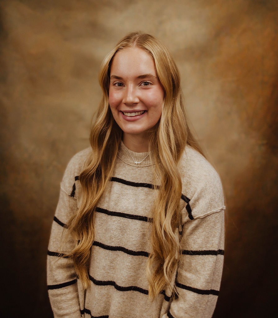 Ella, with her long blonde hair, smiles at the camera while wearing a beige sweater with black stripes. The backdrop is a warm, textured brown, enhancing her radiant presence.