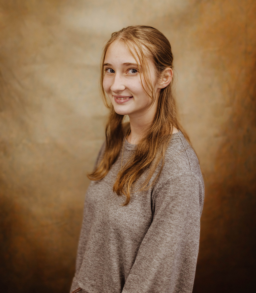 A young woman with long hair, named Ellie, smiles softly at the camera. She is wearing a gray sweater and stands against a warm, textured brown background.