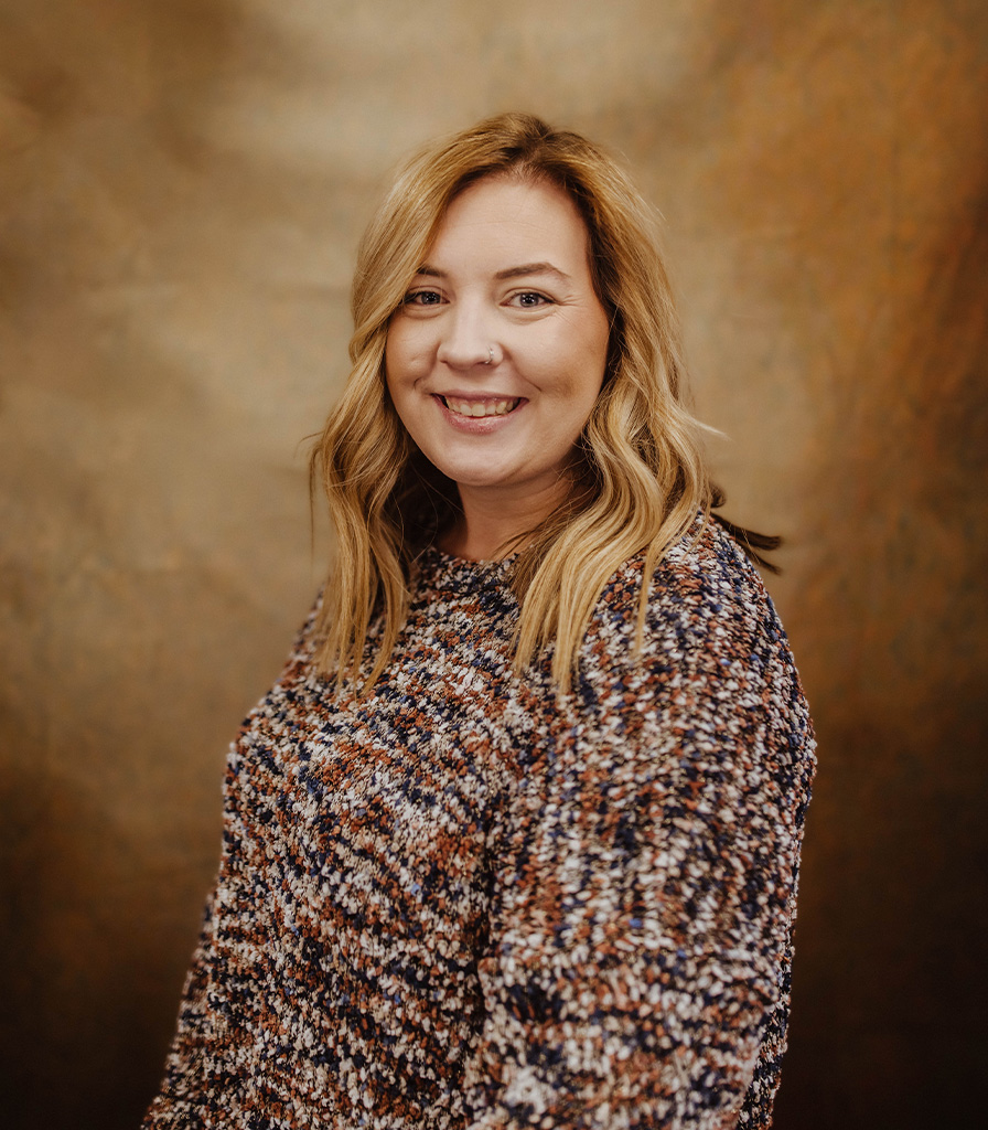 Haley, with her shoulder-length blonde hair, smiles warmly while wearing a speckled sweater, posing against a rich, textured backdrop.