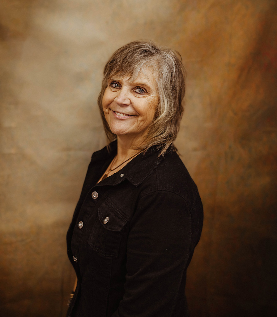 Jan, with shoulder-length hair, smiles at the camera while wearing a dark jacket against a brown, textured background.