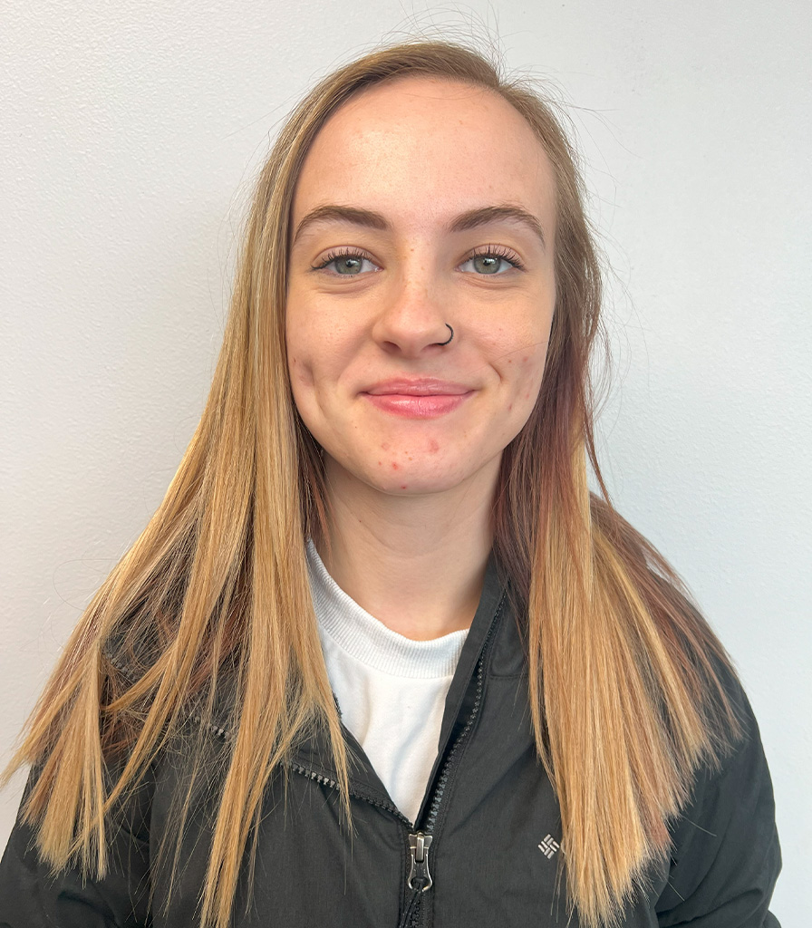 Kallie, with long, straight blonde hair, smiles warmly at the camera against a plain white background. She's dressed in a black jacket over a white t-shirt and sports a stylish nose ring.