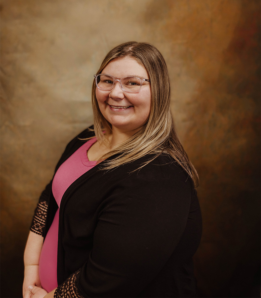 Katie, a woman with long blonde hair and glasses, smiles warmly in her pink top and black cardigan. She stands gracefully against a brown textured background.