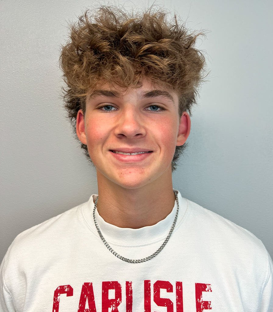 Kyler, a young person with curly brown hair, smiles at the camera. They are wearing a white T-shirt with red lettering and a silver chain necklace, standing against a plain light gray wall.