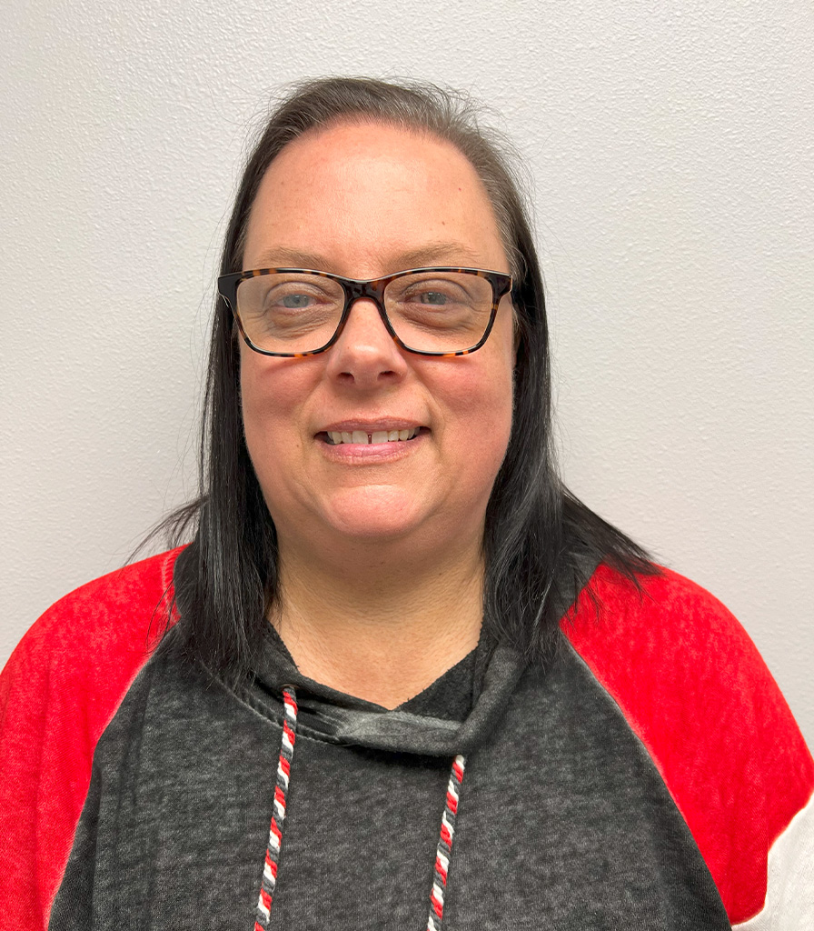 Mary, with shoulder-length dark hair, wearing glasses and a black and red hoodie, smiles warmly in front of a plain white wall.