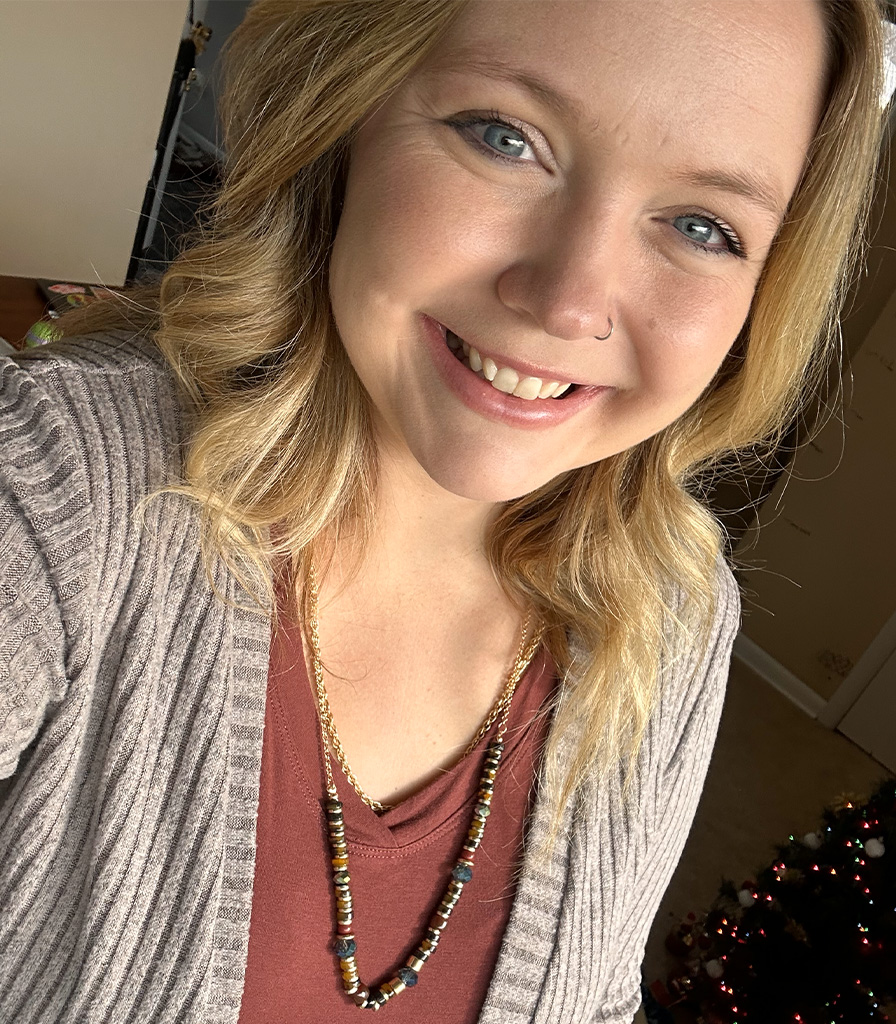 Miranda with blonde hair smiles while taking a selfie indoors. She is wearing a brown top, a textured cardigan, and a long beaded necklace. In the background, there is a small decorated Christmas tree.