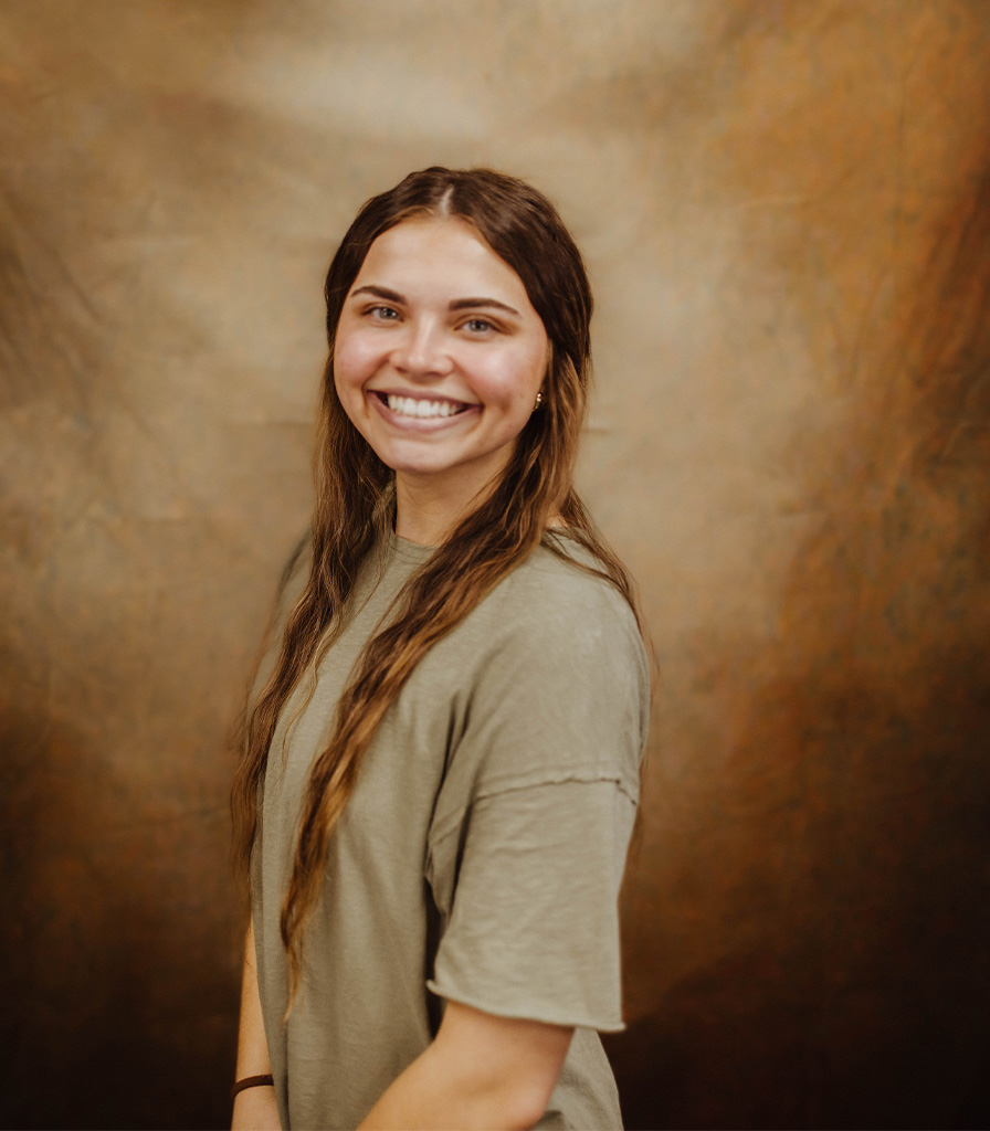 Natalea, a young woman with long brown hair, smiles while standing against a warm, textured brown background. She is wearing a short-sleeved, light olive-green top.