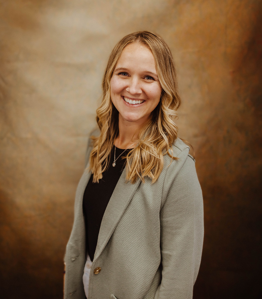 Paige, a woman with long blonde hair, smiles warmly at the camera. She is wearing a light gray blazer over a black top. The background is a textured, warm brown color.