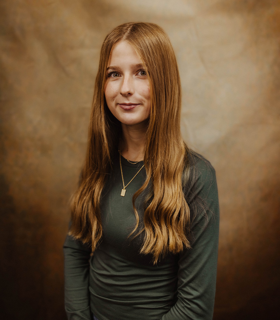 A young woman named Parker, with long, wavy hair and a pendant necklace, stands against a textured brown backdrop wearing a dark green, long-sleeved shirt.