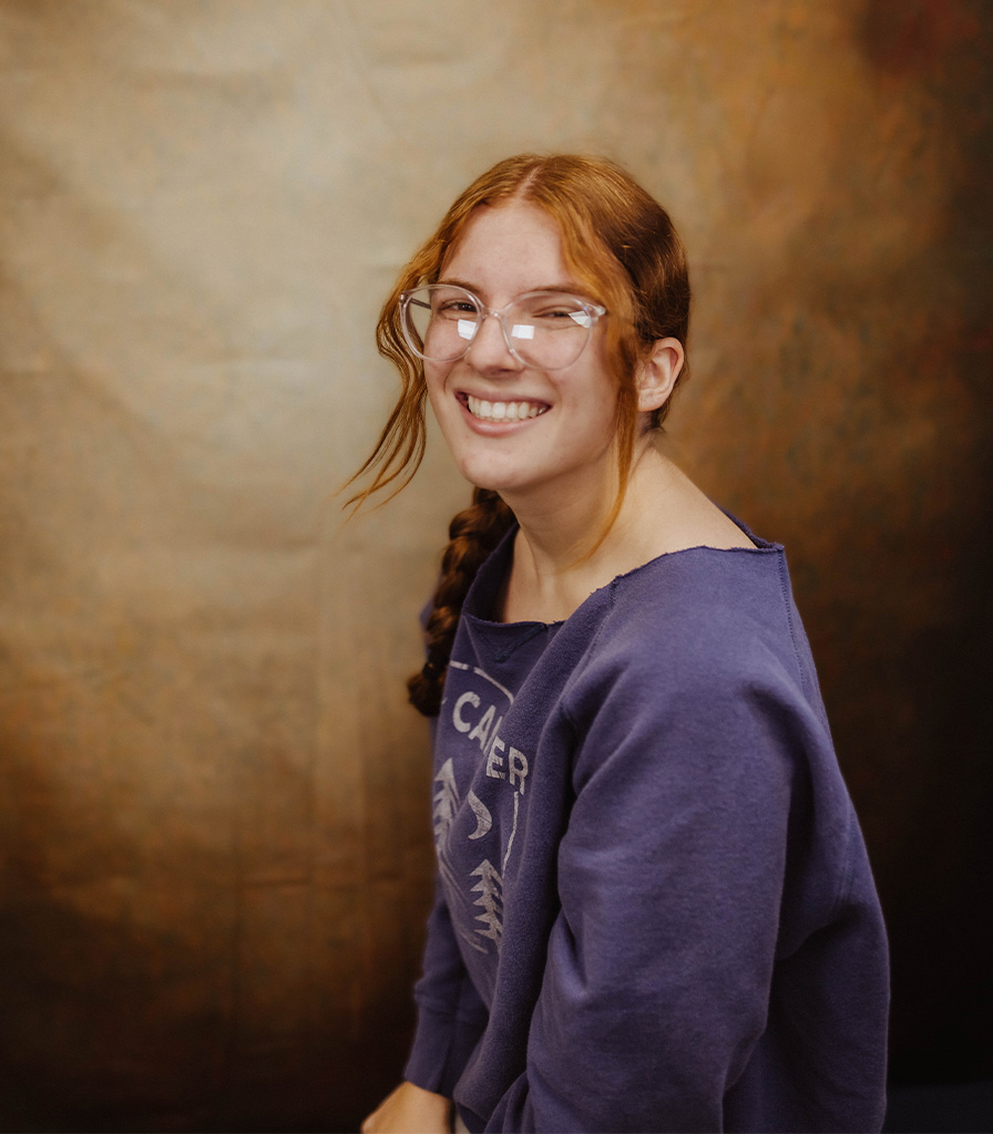 Sam, with long red hair and glasses, smiles warmly while wearing a purple sweatshirt against a softly blurred brown background.