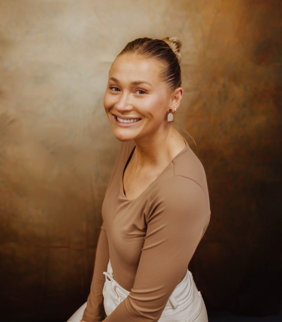 Tristine, with light hair tied up and wearing a tan top, smiles at the camera. The background is a warm, textured brown hue.