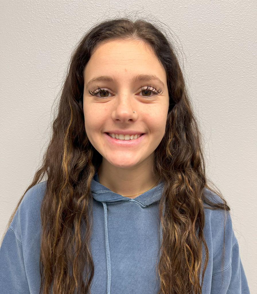 Zaddie with long wavy brown hair is smiling at the camera. They are wearing a blue hoodie against a plain white background.